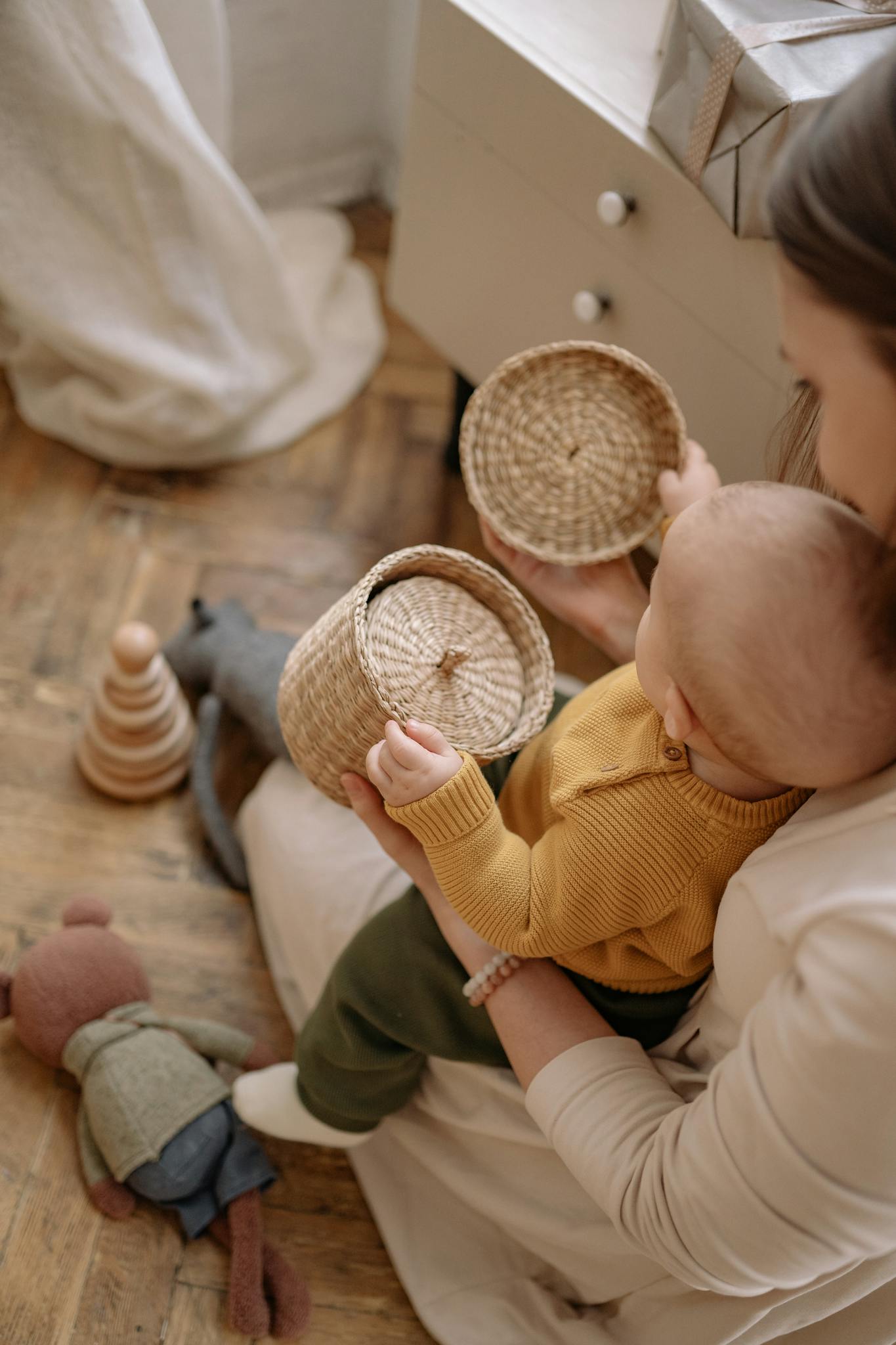 A mother and baby bond while playing with wooden and woven toys on a wooden floor, creating a warm family moment.